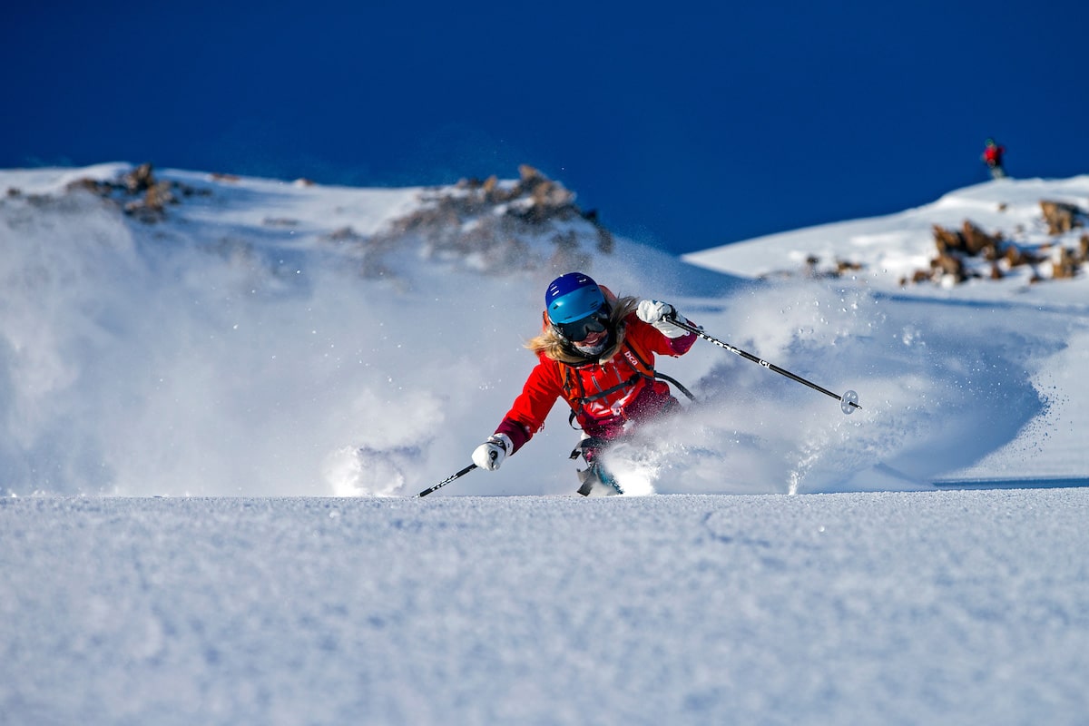 A blonde woman wearing a red snowsuit quickly skis on fresh snow in Alaska.