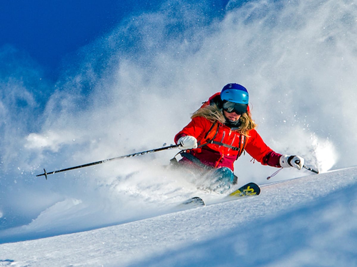 Woman with blonde hair wears a red snowsuit and heli skis down a mountain in Alaska.