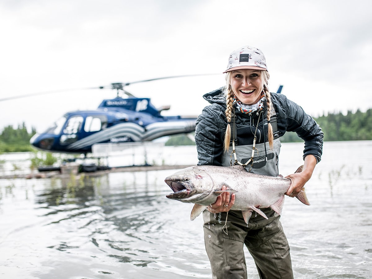 A woman holds a fish she caught during a day of heli fishing with a helicopter behind her.