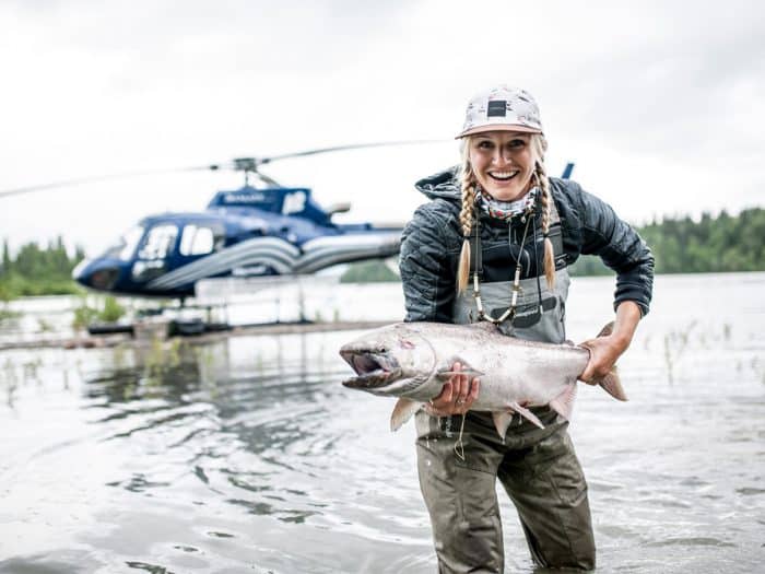 A woman wearing fishing gear smiles as she holds an Alaskan salmon she just caught.