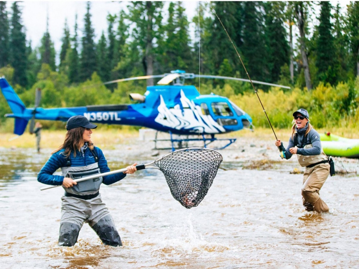 Two people fly fish in a river near Tordrillo Mountain Lodge with a helicopter in the background.