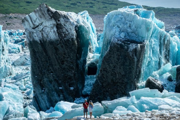 Two people stand between a cracked glacier with white and bright blue ice in Alaska.