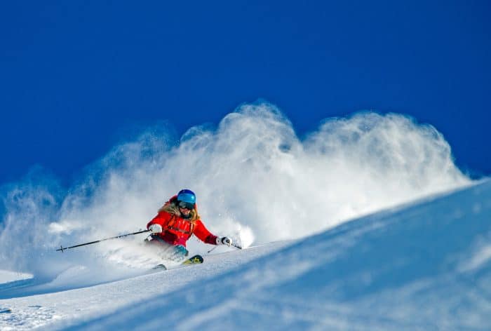 Female skier in a red coat and goggles skis down a mountain of fresh powder.