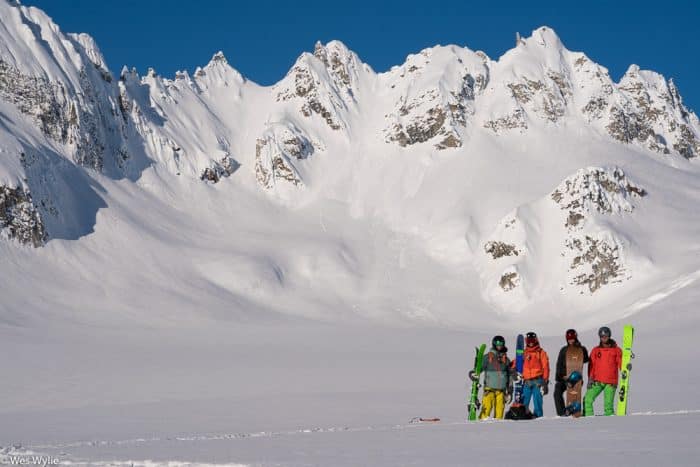 A group of four heli skiers and heli snowboarders pose with a large snow-covered mountain in Alaska.