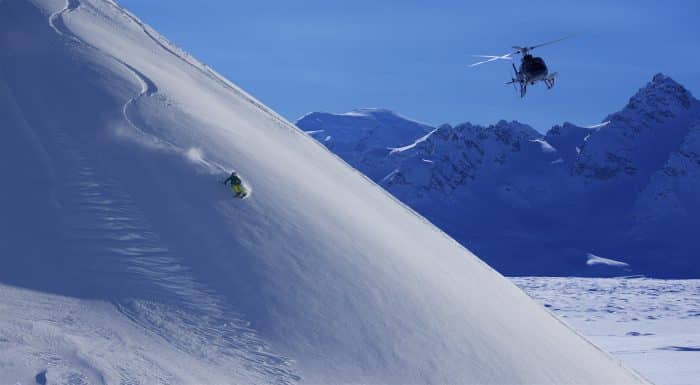 A heli skier makes a run through fresh powder as a helicopter hovers nearby at Tordrillo Mountain Lodge.