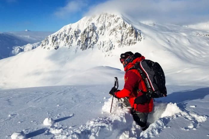 Skier in red coat and black backpack with snow covered mountain in the background.