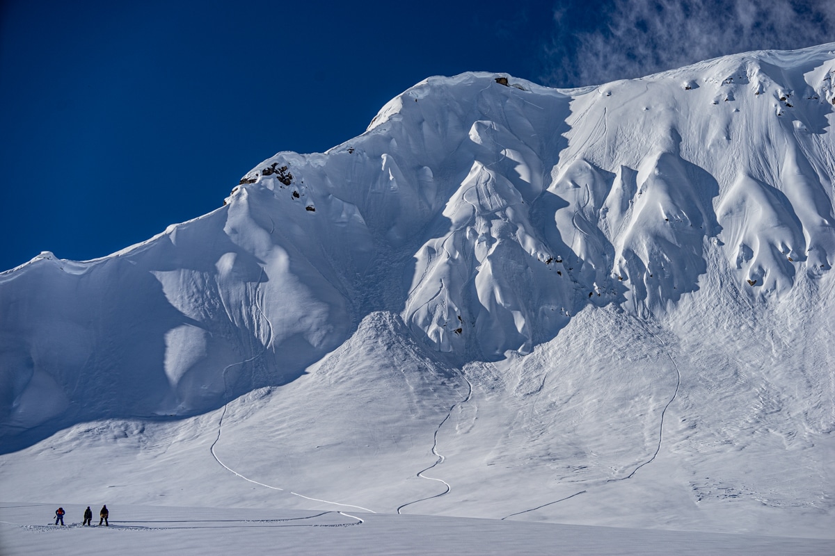 Group of three skiers and snowboarders at the bottom of a snowy mountain in Alaska.