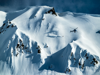 Black helicopter flies over snow capped Tordrillo Mountains in Alaska as a skier glides down.