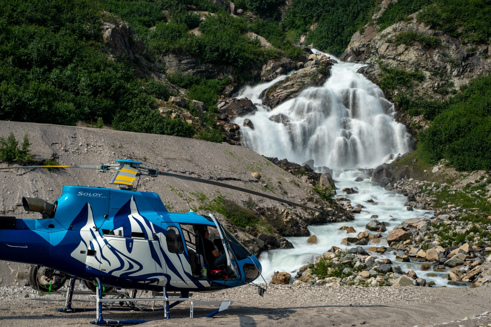 a helicopter and its pilot sits under a waterfall in the tordrillo mountains