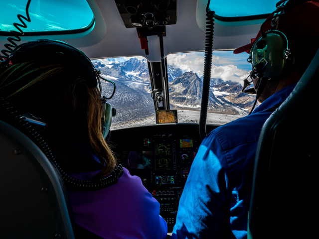 a passenger and pilot fly in a helicopter above the triumvirate glacier